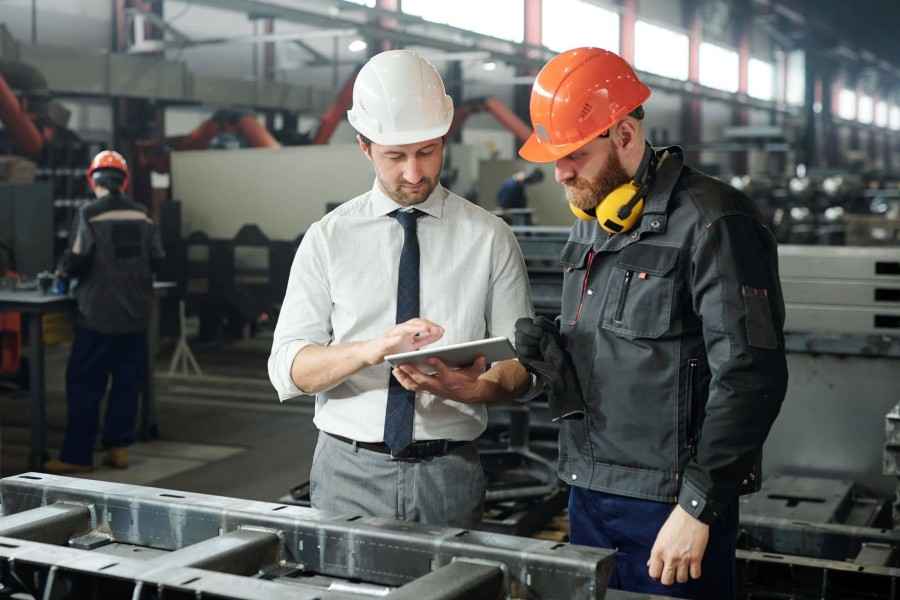 Fokusbranche Maschinenbau und Metallverarbeitung, Baustein Sachversicherung: 2 Mitarbeitende mit Helm in Fabrikhalle konfigurieren eine Maschine.