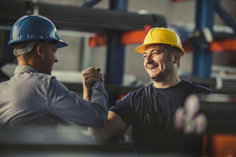 Fokusbranche Maschinenbau und Metallverarbeitung, Zusatzbaustein Gruppen-Unfallversicherung: Zwei Mitarbeitender grüßen sich in einer Lager-/Produktionshalle per Handschlag