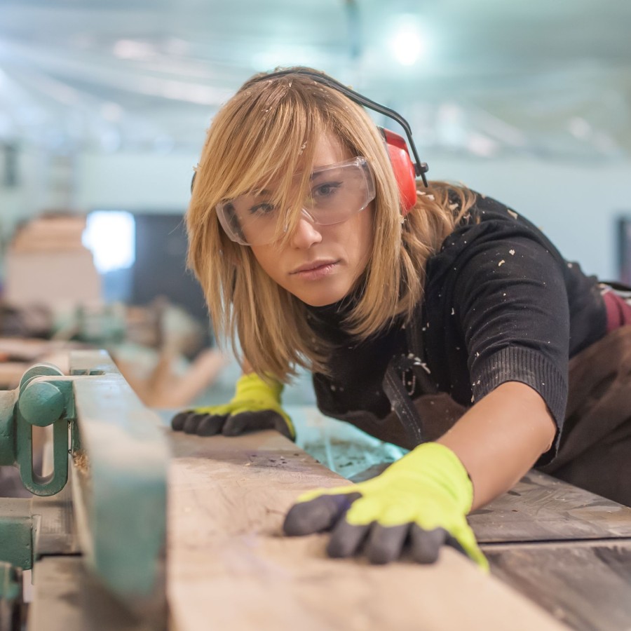 Eine Tischlerin mit Ohren- und Augenschutz sägt Holz an einer elektrischen Maschine zu