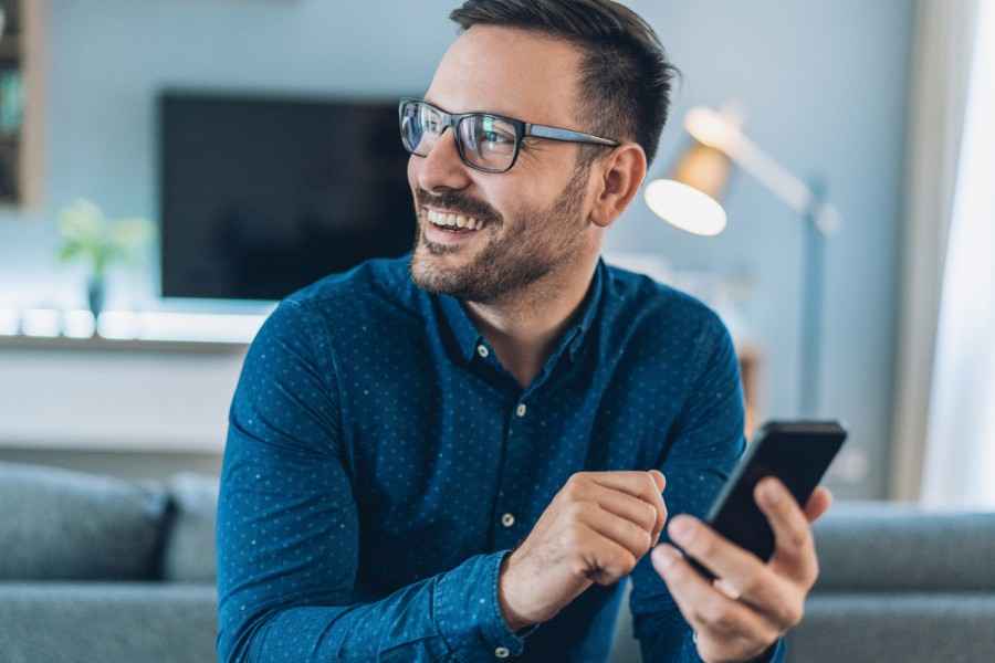 Ein Mann sitzt im Wohnzimmer und hält ein Smartphone in der Hand.