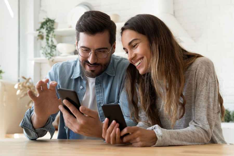 Gesundheitsapp: Junge Frau und junger Mann - beide mit Smartphone - sitzen am Tisch und freuen sich über die App.