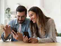 Gesundheitsapp: Junge Frau und junger Mann - beide mit Smartphone - sitzen am Tisch und freuen sich über die App.