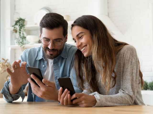 Gesundheitsapp: Junge Frau und junger Mann - beide mit Smartphone - sitzen am Tisch und freuen sich über die App.