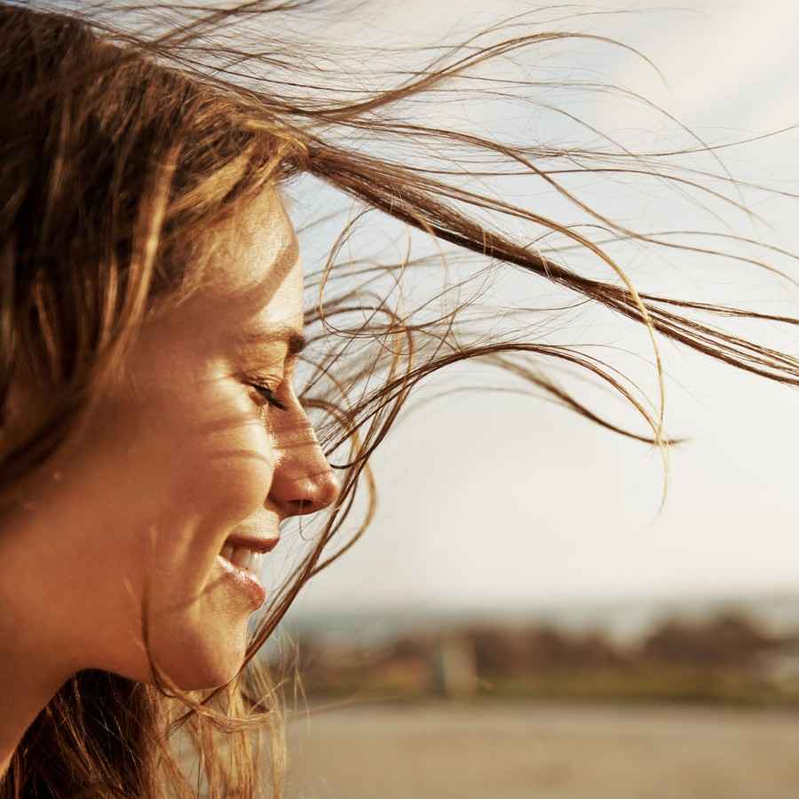 Frau mit wehenden Haaren hält ihr Gesicht mit geschlossenen Augen in die Sonne.