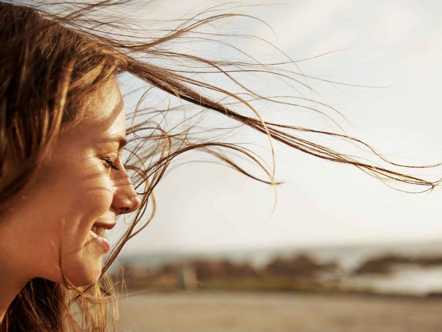 Frau mit wehenden Haaren hält ihr Gesicht mit geschlossenen Augen in die Sonne.