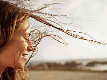 Frau mit wehenden Haaren hält ihr Gesicht mit geschlossenen Augen in die Sonne.