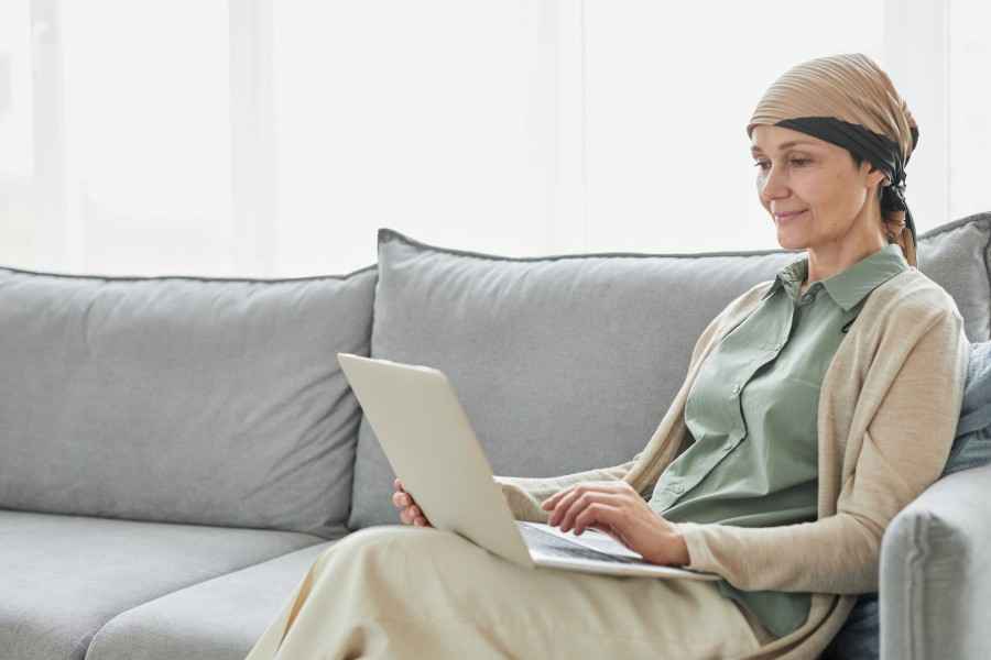 Krebserkrankung - eine Frau mit Kopftuch sitzt auf dem Sofa, einen Laptop auf dem Schoß