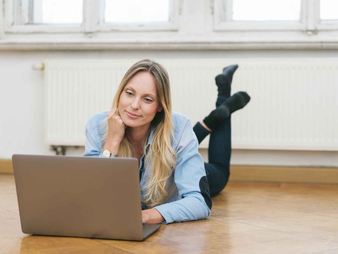 Frau liegt bäuchlings auf ihrem Parkettboden und schaut in ihren Laptop