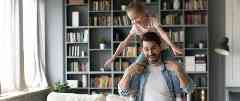 Hausratversicherung: Vater hat Tochter auf seinen Schultern. Sie lachen beide und laufen durch ihr Wohnzimmer.