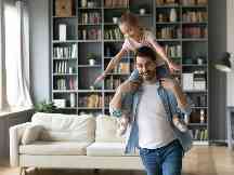 Hausratversicherung: Vater hat Tochter auf seinen Schultern. Sie lachen beide und laufen durch ihr Wohnzimmer.