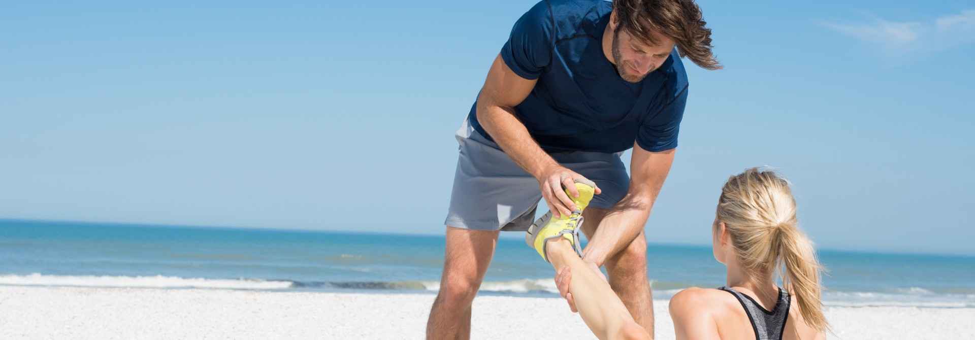 Gothaer Unfallversicherung: Jogger schaut sich am Strand den Fuß seiner Freundin an.