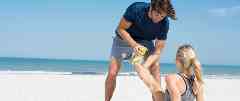 Gothaer Unfallversicherung: Jogger schaut sich am Strand den Fuß seiner Freundin an.