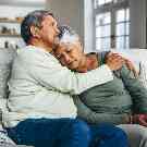 Älteres Ehepaar sitzt zusammen auf der Couch. Der Mann hält seine Frau tröstend im Arm. 