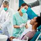 Frau liegt beim Zahnarzt auf einem Behandlungsstuhl. Ihre Zähne werden von einem Zahnarzt und einer Zahnarzthelferin behandelt.