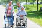 Ehefrau mit Rollator und Ehemann im Rollstuhl gehen mit ihrer Pflegerin im Park spazieren.