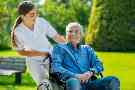 Eine Pflegerin schiebt einen älteren Patienten im Rollstuhl im Park spazieren.