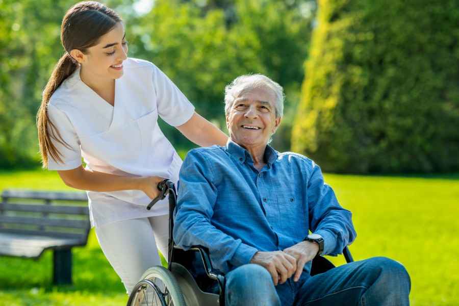Eine Pflegerin schiebt einen älteren Patienten im Rollstuhl im Park spazieren.