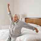 Rentnerin wacht zufrieden auf. Sie sitzt im Schlafanzug auf der Kante ihres Bettes und streckt sich.