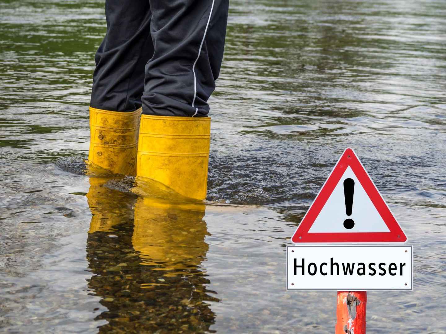 Ausschnitt von Beinen in Gummistiefeln neben Hochwasser-Schild im Hochwasser