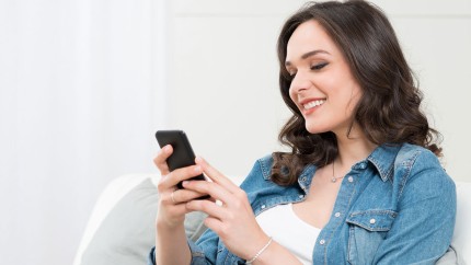 GesundheitsApp: Junge Frau sitzt mit dem Smartphone auf dem Sofa und lädt sich die App herunter. GesundheitsApp: Junge Frau sitzt mit dem Smartphone auf dem Sofa und lädt sich die App herunter.