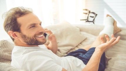 Informationen für Krankenversicherte: Junger Mann, der ein Bein gebrochen hat, auf der Couch am telefonieren. Informationen für Krankenversicherte: Junger Mann, der ein Bein gebrochen hat, auf der Couch am telefonieren.