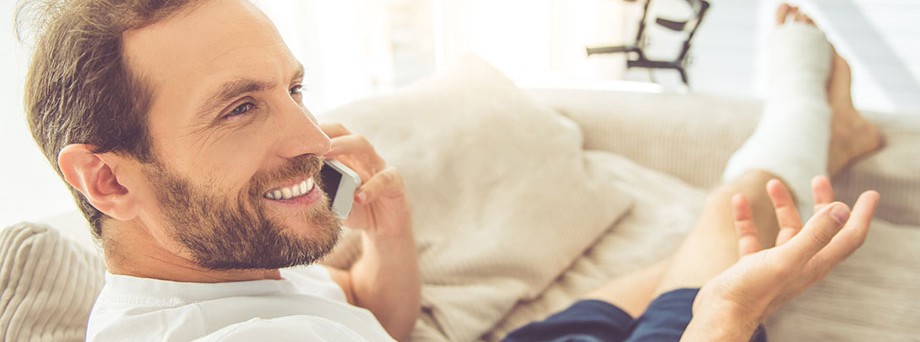 Informationen für Krankenversicherte: Junger Mann, der ein Bein gebrochen hat, auf der Couch am telefonieren. Informationen für Krankenversicherte: Junger Mann, der ein Bein gebrochen hat, auf der Couch am telefonieren.